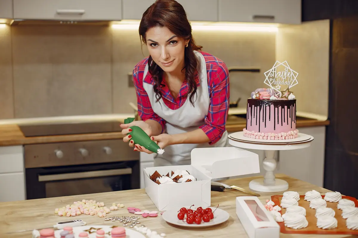 A woman in an apron decorates a cake with icing in a contemporary kitchen setting.