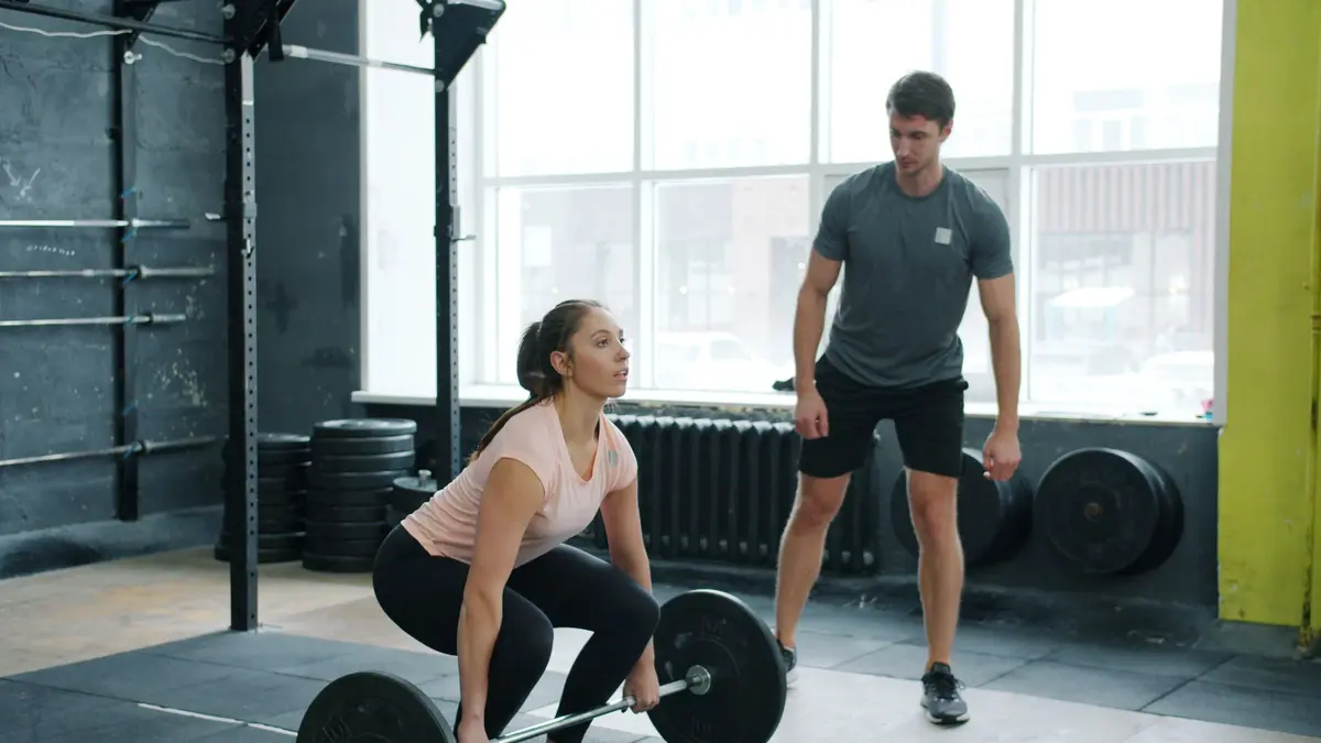 A woman performs deadlifts with a trainer overseeing in a modern gym.