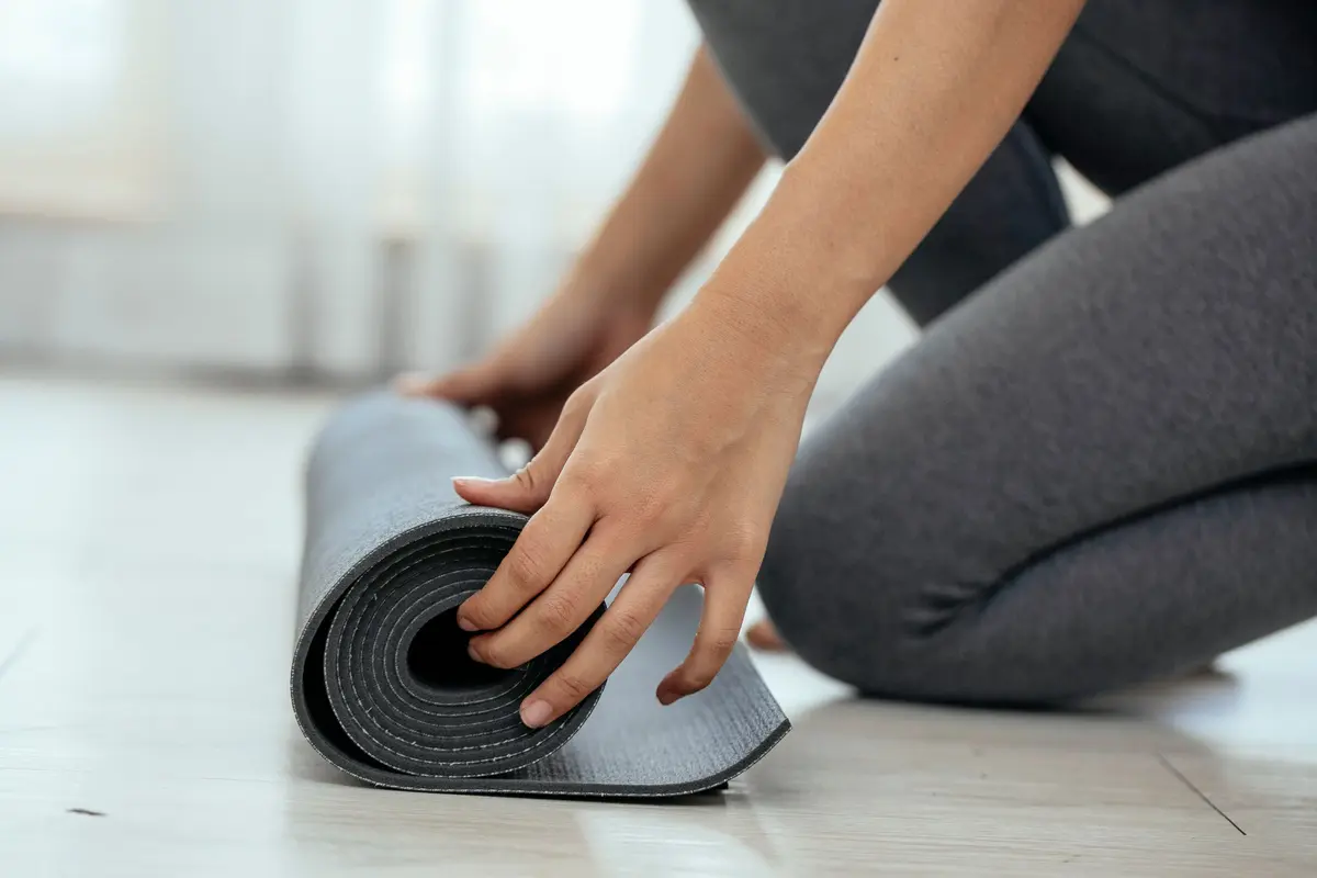 Side view of crop unrecognizable female sitting on floor and unrolling mat in light gym